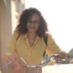 From below cheerful young curly woman in glasses with backpack smiling at camera while riding bicycle along street in sunny day