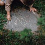High angle of unrecognizable child drawing with chalks on small piece of concrete block in grassy yard