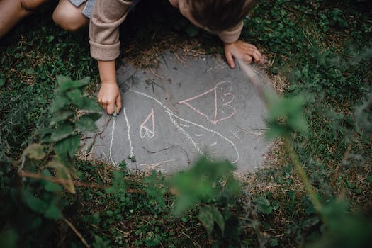 High angle of unrecognizable child drawing with chalks on small piece of concrete block in grassy yard