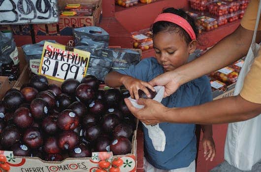 Positive Asian girl putting ripe plums into bag while buying fruits with mother in local street bazaar