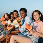 Young content multiracial friends enjoying sweet ripe watermelon slices on sandy sea shore while looking at camera