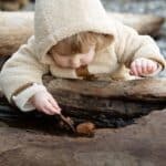 Cute little girl in warm coat playing with small pebbles on wet log while resting on shore in autumn day