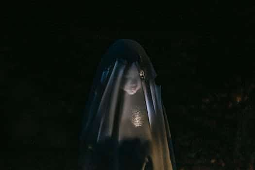 Mysterious little girl standing in darkness covered with white blanket as ghost and shining flashlight on face