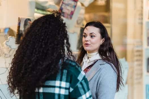 Positive woman communicating with unrecognizable female friend while standing near glass shop window of store with souvenirs on street in city