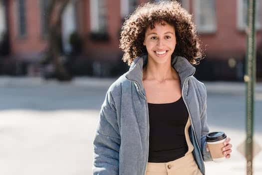 Confident young ethnic female millennial with curly dark hair in trendy outfit smiling and looking away while standing on city street with cup of takeaway coffee in hand