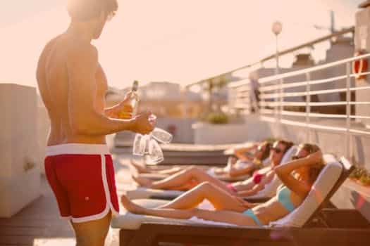 Side view of handsome young muscular guy holding bottle and glasses and offering drink to women sunbathing on lounges while resting together on sunny terrace