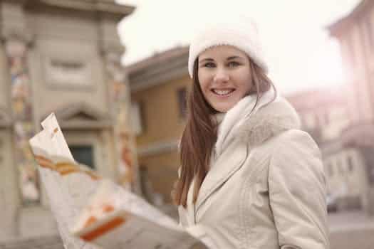 Young female tourist in warm jacket hat and scarf holding paper map and looking at camera while standing in historical center of city