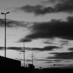 Low angle of black and white distant car with headlights riding on dark rural road near fences and streetlights against cloudy sky in evening