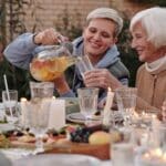 Positive lady pouring drink into glass for elderly woman while having dinner with family on terrace