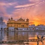 Exterior of Sikh gurdwara golden temple with dome located near water against cloudy sky in evening time in city in India