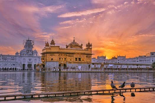 Exterior of Sikh gurdwara golden temple with dome located near water against cloudy sky in evening time in city in India