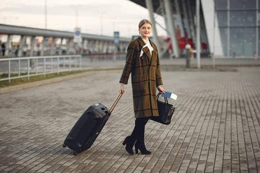 Full body young female passenger in trendy plaid coat with suitcase strolling on pavement near modern building of ariport terminal taking bag and documents