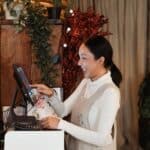 Side view of smiling young Asian female cashier wearing apron using modern cash desk with computer screen in cafe with rustic wooden interior