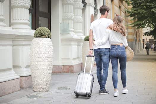 Back view of traveling couple in love wearing casual clothes walking with luggage and hugging while strolling along sidewalk together during vacation