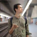 Young ethnic man in earbuds listening to music while waiting for transport at contemporary subway station