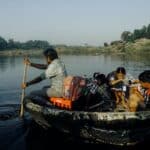 Group of Indian man and schoolchildren with backpacks sitting in handmade boat while floating on water to school